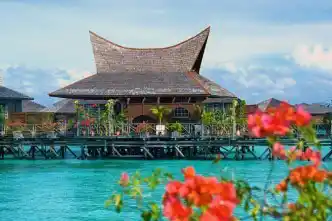Iconic architecture of a Borneo Villa Suite at Mabul Water Bungalows, viewed through vibrant red bougainvillea flowers over the turquoise sea.