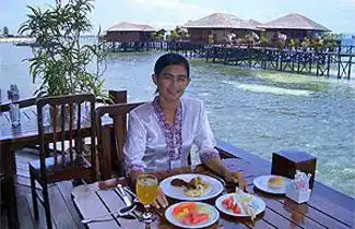 Guest enjoying a delicious meal on the open-air dining deck at Sipadan Water Village, with the resort's overwater bungalows in the background.