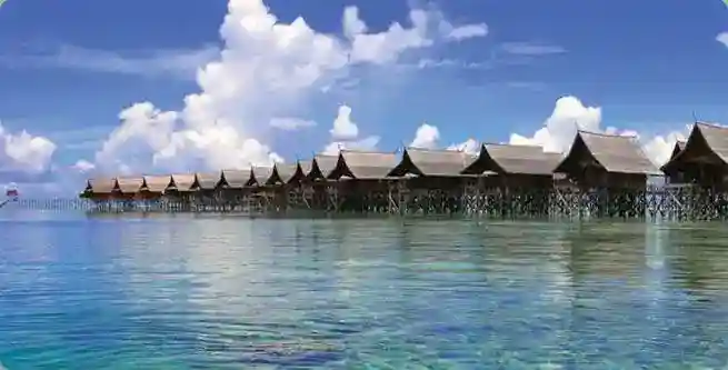Iconic overwater chalets of the Kapalai Dive Resort lined up perfectly over the calm, crystal-clear turquoise sea under a blue sky with fluffy clouds.