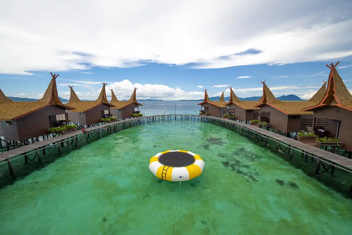 Symmetrical view of Dayang Resort's overwater chalets with their distinctive high-peaked roofs, surrounding a water trampoline in a clear turquoise lagoon.