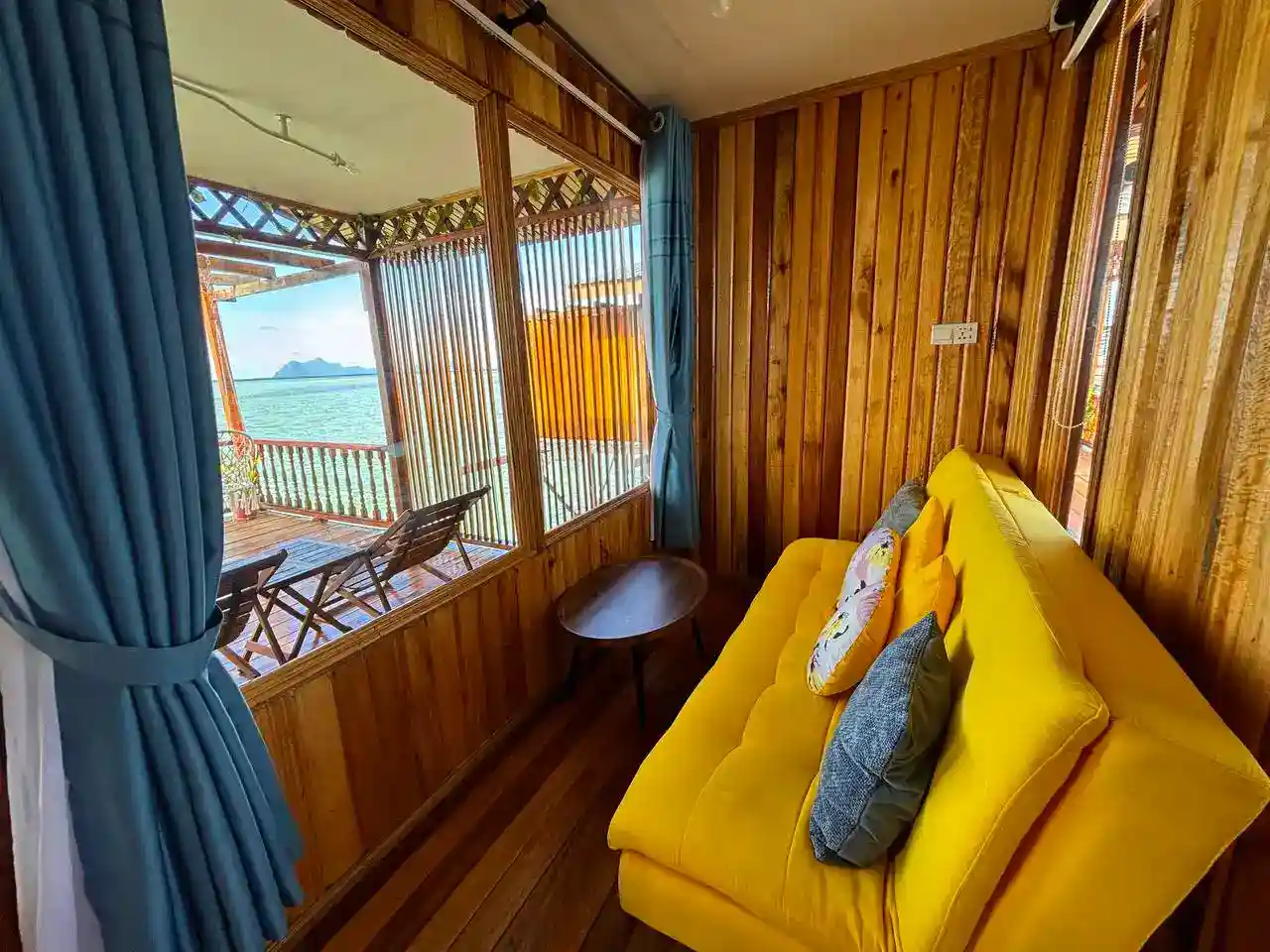 Cozy bedroom at Larapan Water Village featuring a bright yellow sofa and large windows that look out onto a private balcony and the sea.
