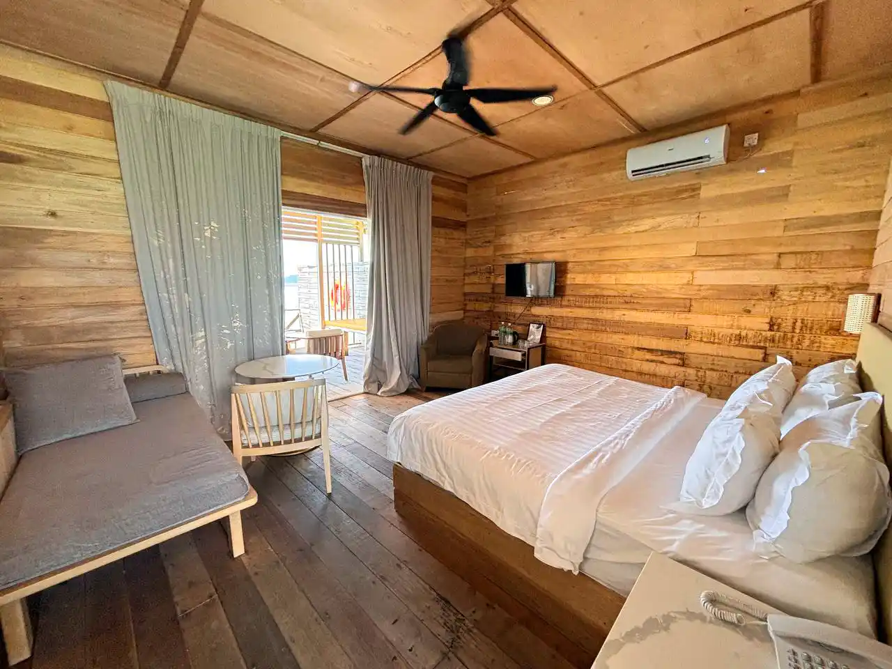 Spacious, wood-paneled bedroom at Sisipan Island Resort, featuring a large bed, a daybed, and an open doorway to the private sea-view balcony.