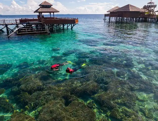 Two snorkelers exploring the vibrant, shallow coral reef directly in front of the Sipadan Water Village overwater bungalows.